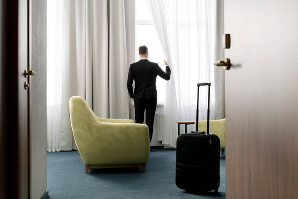 ホテルの部屋に立って暗いスーツを身に着けているビジネスマンの背面図窓を見て - 写真・画像