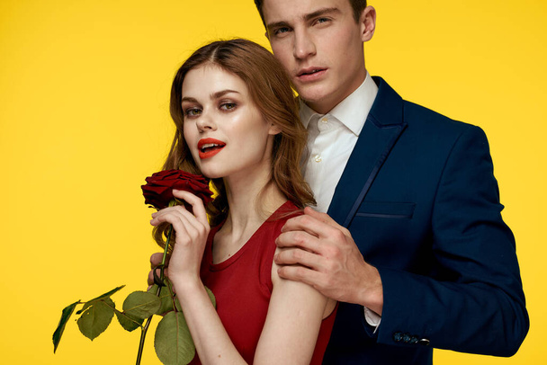 Egy romantikus férfi megölel egy vörös ruhás nőt egy rózsával a kezében, sárga alapon. - Fotó, kép
