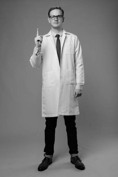 Studio shot of young handsome man doctor with blond hair against gray background in black and white - Foto, Imagen