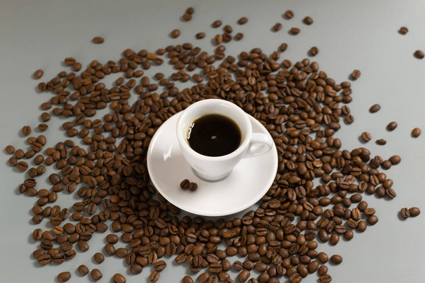 Valkoinen kuppi espresso valkoisella lautasella keskuudessa kahvipapuja harmaalla pohjalla. Lähemmäs. Valikoiva keskittyminen. Vaakasuuntainen. - Valokuva, kuva