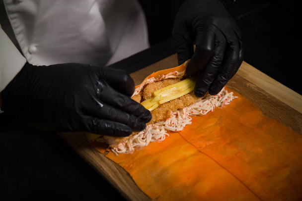 Koken proces van het inpakken van Japanse geen rijst sushi roll in wortel daikon. Chef-kok bereidt Pan Aziatische schotel op houten bord. Restaurant eten maken. Sushi rollen zonder rijst in zwarte beschermende handschoenen - Foto, afbeelding