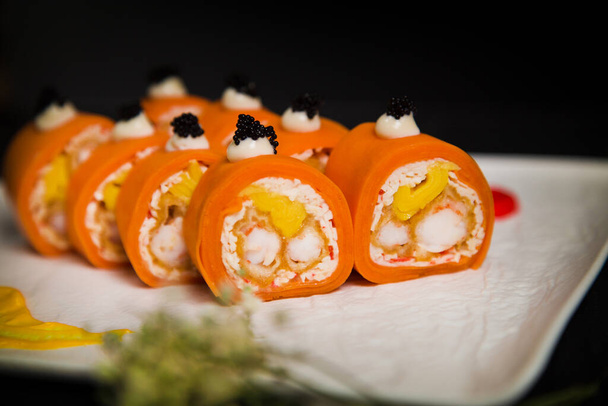 Close up of Asian No Rice Healthy Sushi Roll pieces with tempura shrimp wrapped in carrot daikon served on white Japanese plate with flowers and paining brush strokes. Dark background - Photo, Image