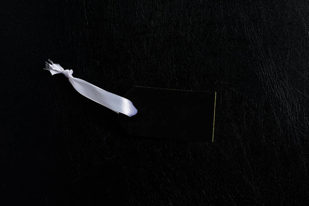 Mockup zwart papieren kledinglabel met wit lint op een donkere eco lederen achtergrond, kopieer ruimte. Bovenaanzicht. Black Friday Sale concept. - Foto, afbeelding