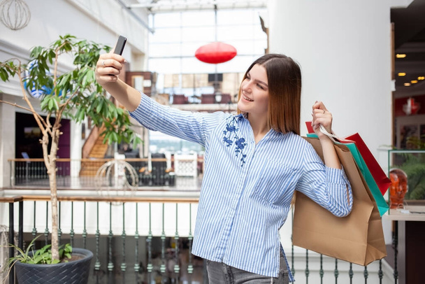 Zdjęcie młodej Europejki, ze szczęśliwą twarzą, stojącej w centrum handlowym z torbami na zakupy i robiącej selfie na smartfonie. Zakupy i koncepcja sprzedaży. Wewnątrz, wewnątrz. - Zdjęcie, obraz