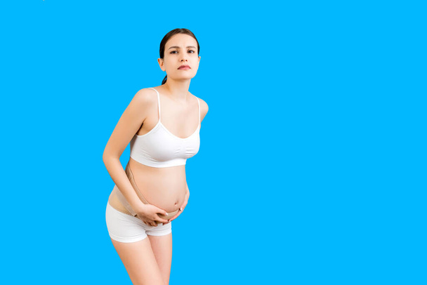 Porträt einer Stützbinde gegen Rückenschmerzen bei Schwangeren in Unterwäsche auf blauem Hintergrund mit Kopierraum. Die Mutter leidet unter Rückenschmerzen. Orthopädisches Bauchstützgurt-Konzept. - Foto, Bild