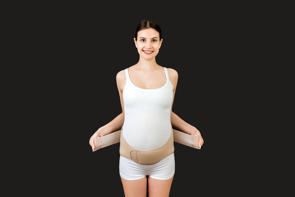 Ritratto di donna incinta che indossa una benda di sostegno per ridurre il mal di schiena su sfondo nero con spazio per la copia. Cintura di supporto addominale ortopedico concetto. - Foto, immagini