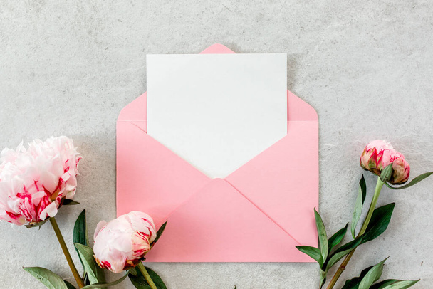 Mockup invitation, blank paper greeting card, pink envelope and peonies on gray stone table. Flower background. Flat lay, top view. - 写真・画像