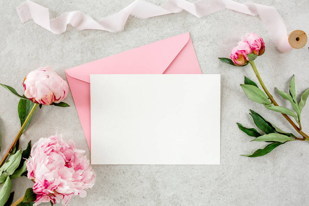 Mockup invitation, blank paper greeting card, pink envelope and peonies on gray stone table. Flower background. Flat lay, top view. - Фото, изображение