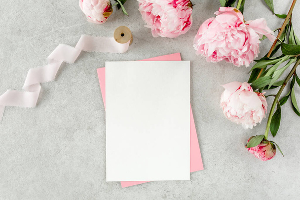 Mockup invitation, blank paper greeting card, pink envelope and peonies on gray stone table. Flower background. Flat lay, top view. - Fotoğraf, Görsel