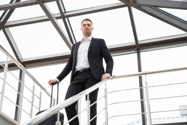 Junger Geschäftsmann in festlichem Anzug steht mit Koffer auf einer Treppe in modernem Gebäude - Foto, Bild