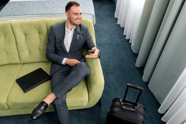 Cheerful businessman using phone sitting in hotel room with suitcase on business trip - Photo, Image
