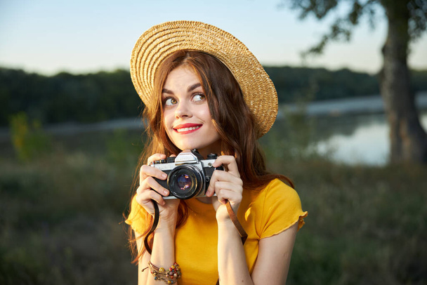 Egy női fotós, akinek kamera van a kezében, oldalra néz, megharapja a száját egy kalapban a természet hátterében. - Fotó, kép