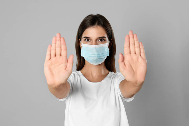 Woman in protective mask showing stop gesture on grey background. Prevent spreading of coronavirus - Foto, immagini