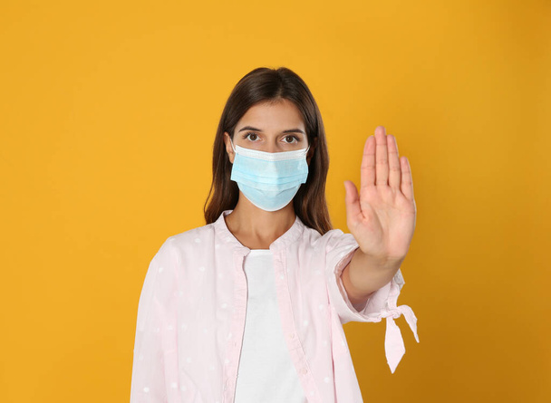 Woman in protective mask showing stop gesture on yellow background. Prevent spreading of coronavirus - Foto, immagini