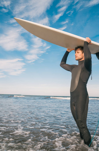 Portrait of young surfer at the beach holding up his surfboard and wearing a black surfing suit. Sport and water sport concept. - Foto, afbeelding