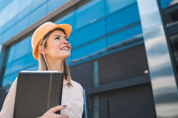 Portrait of professional architect woman wearing yellow helmet and looking at modern building outdoors. Engineer and architect concept. - Foto, afbeelding