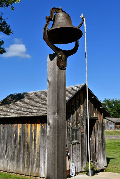 Eine gusseiserne Glocke mit einem Zugseil steht vor einem historischen, restaurierten alten Ein-Zimmer-Schulhaus - Foto, Bild