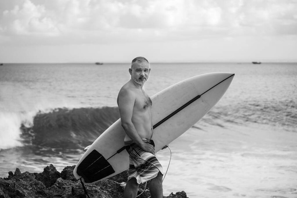 Black and white portrait of handsome shirtless man surfer, holding white surf board  and cactus on background in Bali - Fotó, kép