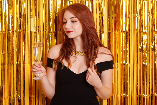 Red haired lady with champagne glass, looking dreamily aside, touching her curlers, posing against yellow wall decorated with glitter, girl wearing black dress. - Фото, изображение