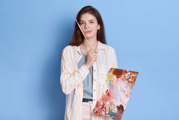 Artist paints picture, posing with pensive expression, looking directly at camera, brunette lady wearing white shirt with brush in hands against blue background. - Fotoğraf, Görsel