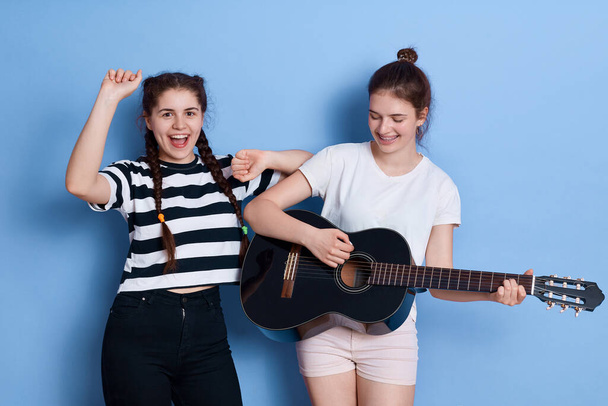 Two friends singing and dancing isolated over blue background, lady with knot playing guitar, winsome girl in striped t shirt and pigtails raising hands up. - Valokuva, kuva