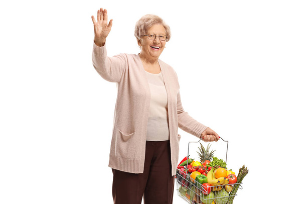 Older woman with fruits and vegetables in a shopping basket waving isolated on white background - Foto, Imagem