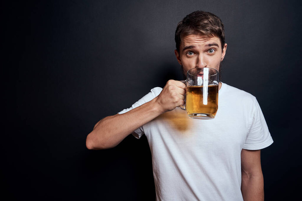 man with a mug of beer in a white t-shirt emotions lifestyle drunk on a dark isolated background - Foto, imagen