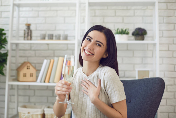 Smiling woman with glass of champagne looking at camera and celebrating holiday online at home - Фото, изображение