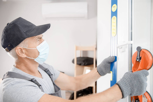Arbeiter in medizinischer Maske montiert Kunststofffenster und -türen mit Staubsauger weiß - Foto, Bild