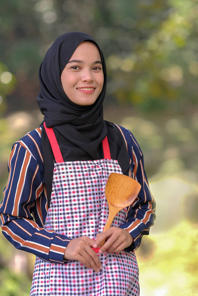 Αναρωτιέται γυναίκα σεφ φορώντας μαντίλα στέκεται χέρι στη μέση κρατά ξύλινη κουτάλα σε ένα θολό φόντο. - Φωτογραφία, εικόνα