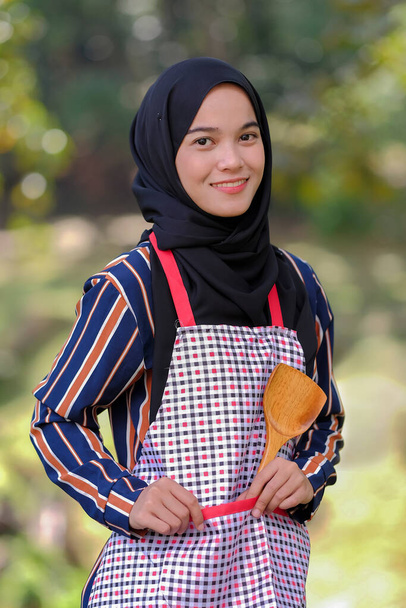 Zastanawiam się kucharka nosząca hidżab stojąca ręka na talii trzyma drewnianą chochlę na rozmytym tle. - Zdjęcie, obraz