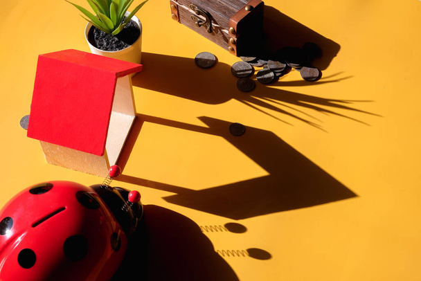 Ladybug κουμπαράς περιβάλλεται από κέρματα και μίνι σπίτι σε κίτρινο φόντο - Φωτογραφία, εικόνα