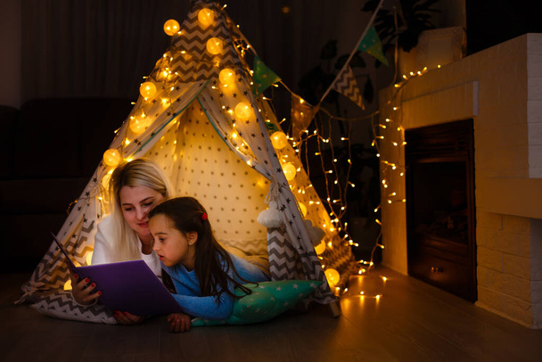 Mère et fille sont assises dans une tente de tipi, lisant des histoires avec la lampe de poche. Famille heureuse. - Photo, image