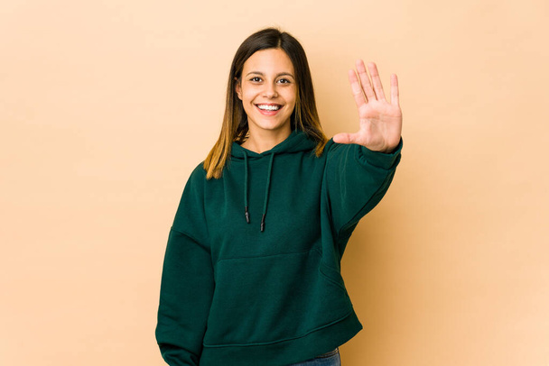 Молодая женщина на бежевом фоне улыбается весело показывая номер пять пальцами. - Фото, изображение