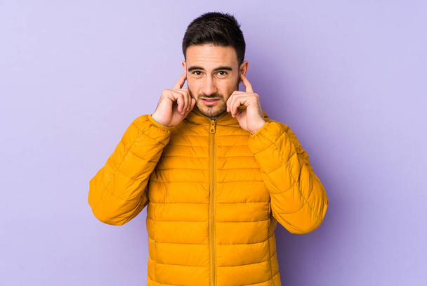 Young caucasian man isolated on purple background covering ears with fingers, stressed and desperate by a loudly ambient. - Photo, Image