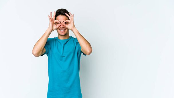 Young caucasian man isolated on white background showing okay sign over eyes - Photo, Image