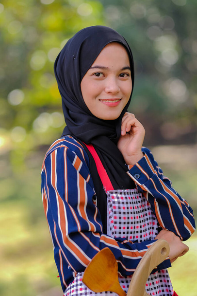Mooie vrouwelijke chef dragen hijab staan met houten lepel en houten snijplank in een schort op een wazige achtergrond. - Foto, afbeelding