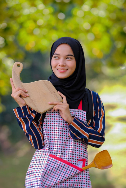 Piękna samica w hidżabie trzymająca drewnianą deskę do krojenia i drewnianą chochlę w fartuchu na rozmytym tle. - Zdjęcie, obraz