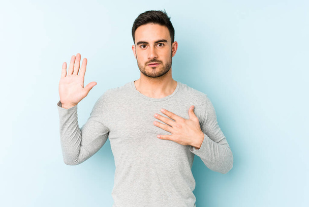 Young caucasian man isolated on blue background taking an oath, putting hand on chest. - Photo, Image