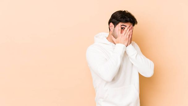 Young man isolated on beige background blink through fingers frightened and nervous. - Photo, Image