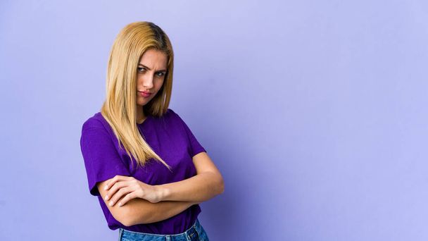 Молодая блондинка изолирована на фиолетовом фоне хмурясь в неудовольствие, держит руки сложенными. - Фото, изображение