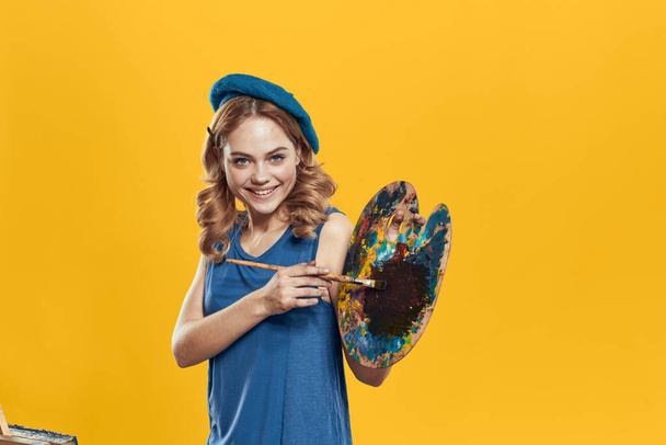 Woman with palette in blue hands takes paintbrush hobby artwork yellow background - Photo, Image