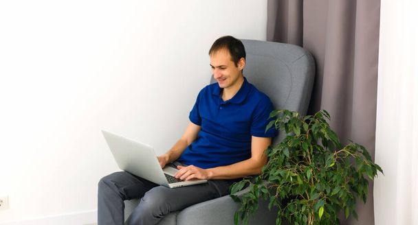Szczęśliwy młody człowiek w t-shircie siedzi na kanapie w domu, pracuje na laptopie, uśmiecha się. - Zdjęcie, obraz