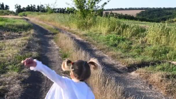 Achterzijde van een klein meisje dat 's zomers wegrent over een onverharde weg op het platteland. Witte cape vliegen in de wind. Blond paardenstaartje fladderend haar. Sereniteit van de kindertijd. Meisje weglopen over landweggetjes midden in zomers landschap - Video