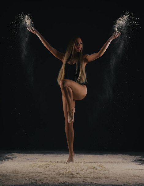 Красивая чувственная стройная танцовщица в черном теле с длинными светлыми волосами бросая пыль, муку, порошок на черном фоне. - Фото, изображение