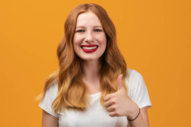 長い波状の赤毛の幸せな女の子は、白いTシャツを着て笑顔と親指を表示します。それは好きよ。いい仕事だ。人間の感情の概念 - 写真・画像