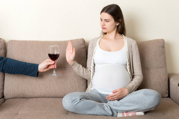 Terhes nő ül a kanapén nem hajlandó bort inni, és megáll gesztus, hogy az üveg. Nincs alkohol a terhesség alatt. - Fotó, kép