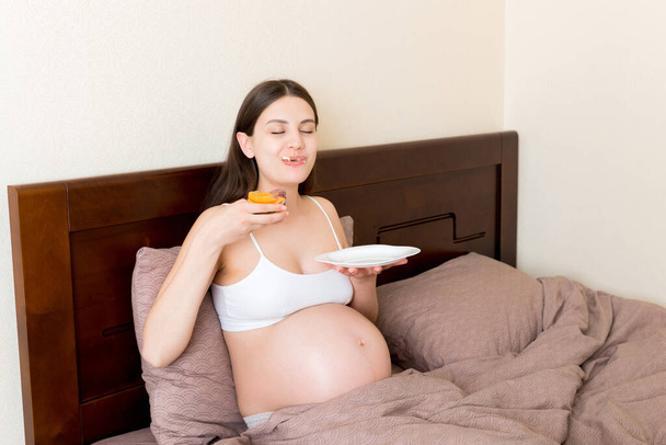 Πεινασμένη έγκυος γυναίκα χαλαρώνοντας στο κρεβάτι τρώει άπληστα ένα κομμάτι κέικ και έχει βρώμικο στόμα. Η μητέρα δεν μπορεί να σταματήσει να τρώει. Έντονη όρεξη κατά τη διάρκεια της εγκυμοσύνης. - Φωτογραφία, εικόνα