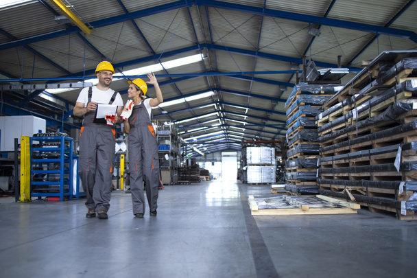 Работники фабрики в рабочей одежде и желтых шлемах, проходящие через производственный цех и обсуждающие вопросы организации. - Фото, изображение