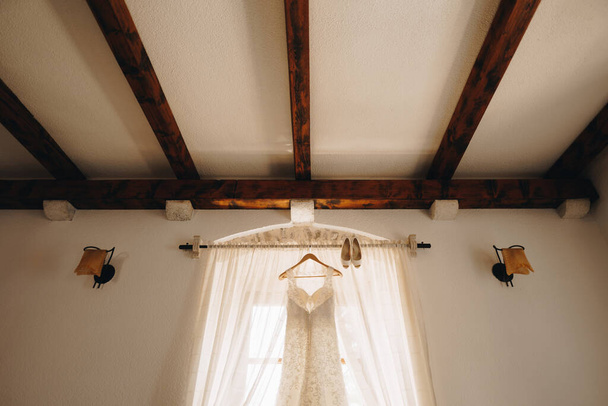 A bright room with a wedding dress on a hanger on the cornice and the brides shoes. - Photo, Image